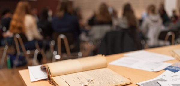 Aufgeschlagene historische Akte im Vordergrund, im Hintergrund sitzen Schülerinnen und Schüler an Tischen.